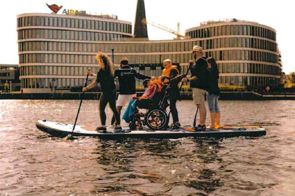 6 Menschen auf einem großen SUP, die Person in der Mitte sitzt im Rollstuhl, gemeinsam genießen sie das paddeln im Rostocker Stadthafen vor dem AIDA-Gebäude